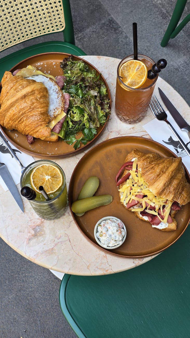 Two croissant sandwiches with drinks and side salad on a cafe table outdoors.