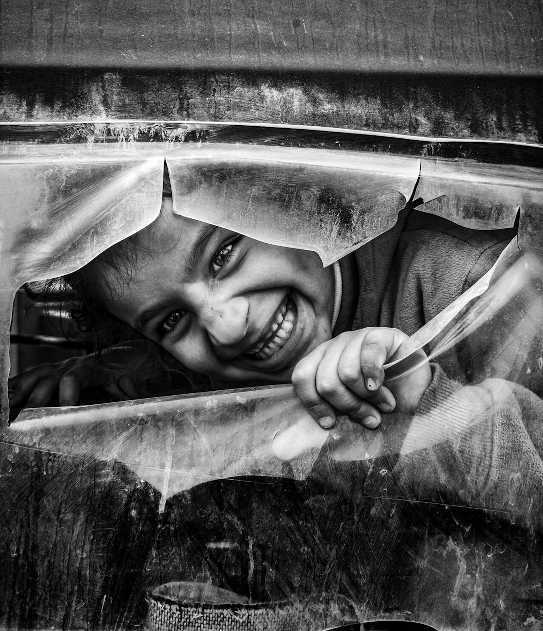 Black and white image of a smiling child peering through a broken window.