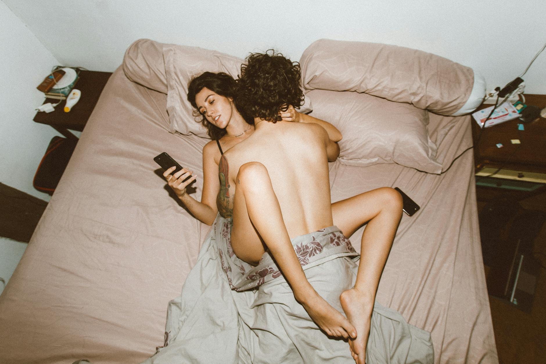 From above of naked young couple having sex on bed in narrow hotel room and browsing mobile phone gadget addiction concept