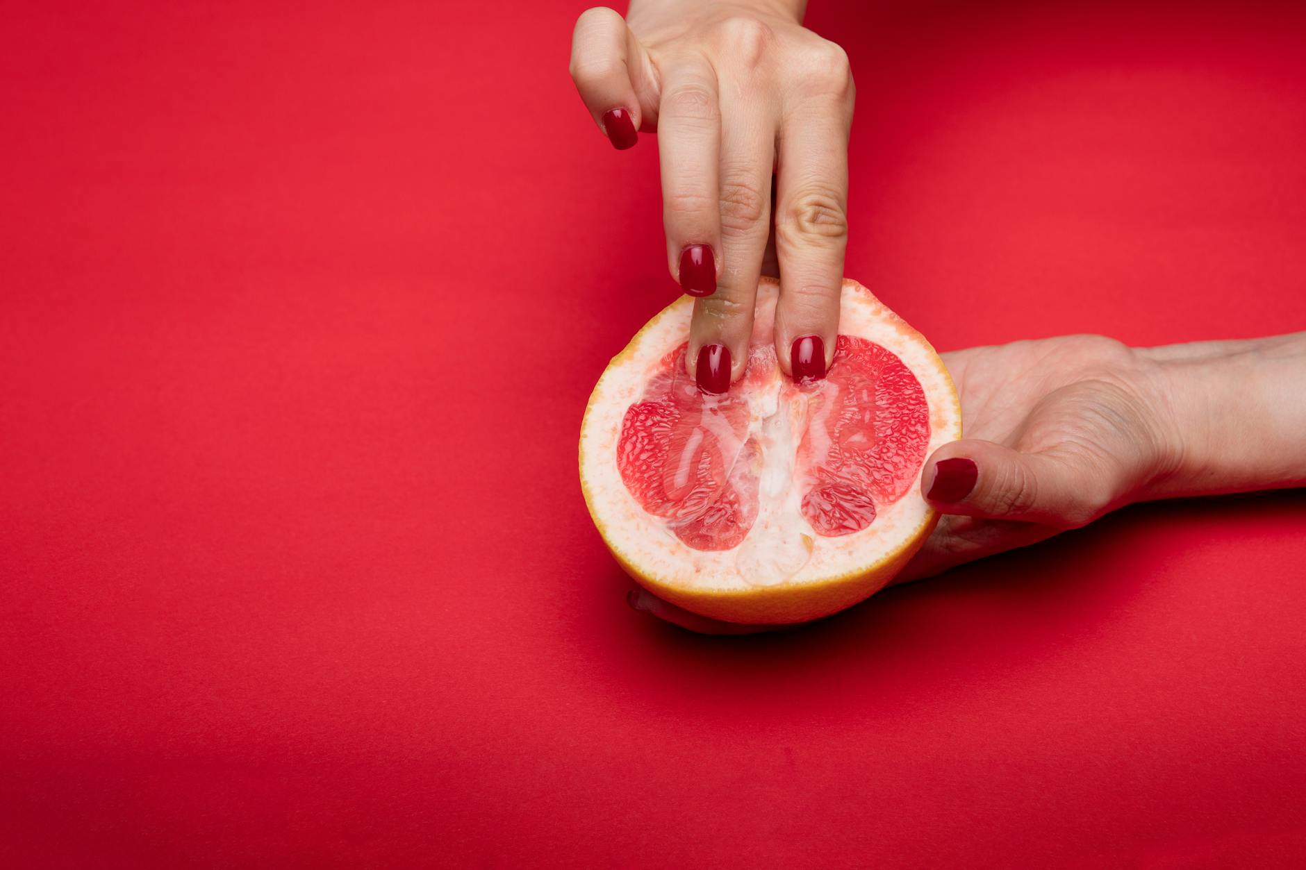 Close-up of hands with red nails holding a halved grapefruit on a red background, conceptual and vibrant.