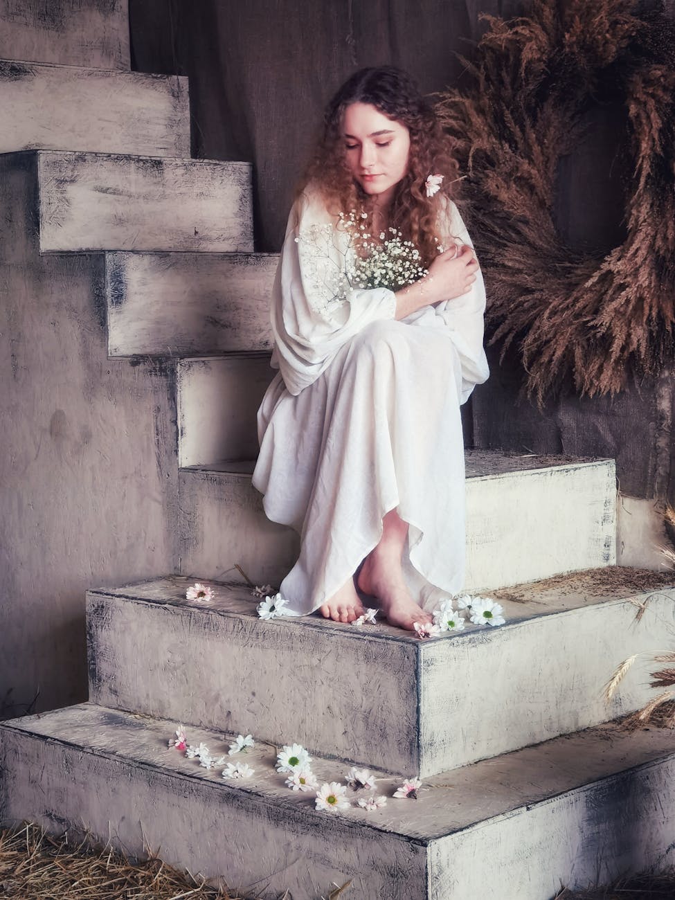 A woman in a white dress sits gracefully on rustic stairs, surrounded by flowers and natural decor.