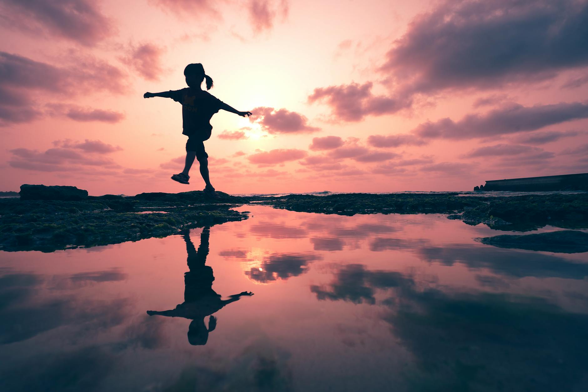 A child silhouette plays near water with a stunning sunset and reflection, creating a serene scene.