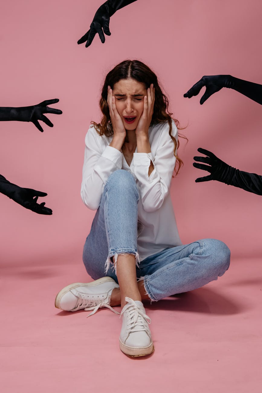 A young woman sits frightened as dark gloved hands reach towards her against a pink background.