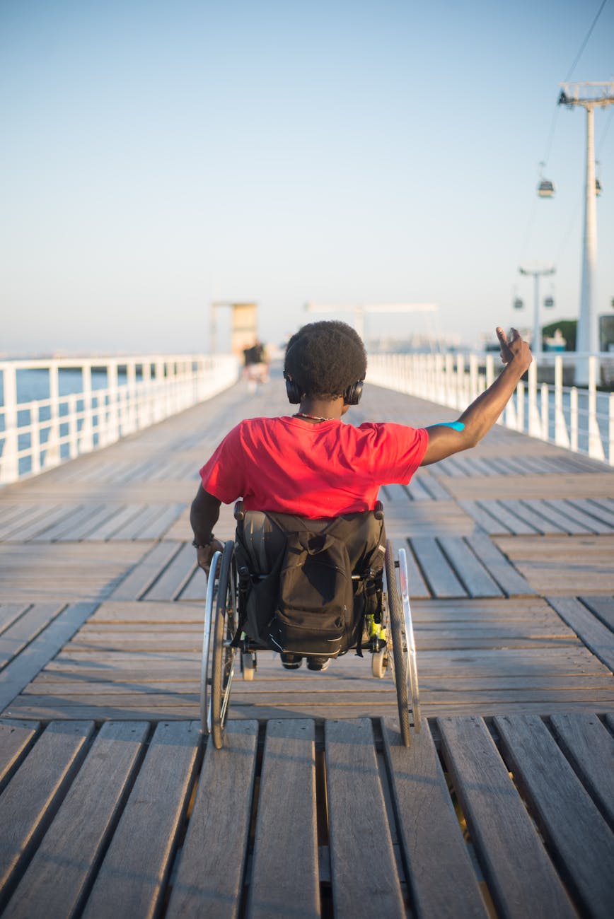 A man in a wheelchair raises his arm while navigating a boardwalk at sunset, symbolizing freedom and positivity.